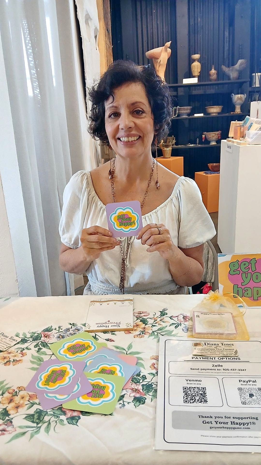 Diana Tenes seated at her Get Your Happy!® table holding a card while offering readings at the Vintage Bazaar Faire inside Mare Island Art Studios and Gallery.